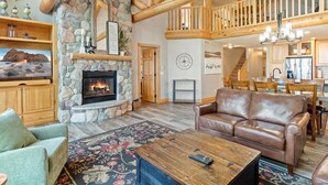 A spacious living room with a stone fireplace, wooden beams, a leather sofa, and a dining area. - Lakefront Townhome | 6BR | Man Cave + Lake View (Nisswa)