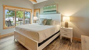A bedroom with a large bed, a ceiling fan, a window with a view of outdoor furniture, and a painting on the wall. - Lakefront Townhome | 6BR | Man Cave + Lake View (Nisswa)