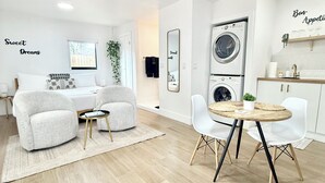 A modern, well-lit room with a bed, two armchairs, a small round table, and a washing machine.
