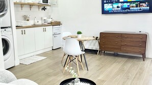 A modern kitchen with white cabinets, a microwave, and a coffee machine. There's a dining area with a table and chairs, and a TV displaying a cooking show.