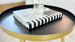 A small round table with a black and gold design, holding a book and a clear vase with white flowers.