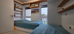 Ett sovrum med två sängar, inbyggda hyllor och ett fönster med utsikt över staden.