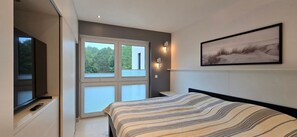 Una camera da letto con una grande finestra, un letto con copriletto a righe, una TV a parete e un quadro incorniciato.