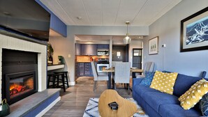 A modern living room with a fireplace, a blue sofa, and a dining area in the background.