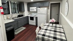 A compact kitchen with grey cabinets, a white microwave, a white refrigerator, a stove, and a sink. There's a small trash bin and a checkered table with chairs. - Cozy Downstairs Apartment (Uniontown)