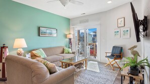 Un salon avec un mur de couleur turquoise, un ventilateur de plafond, un canapé, une table basse, un fauteuil, une lampe et une vue sur la plage, visible à travers une porte vitrée.