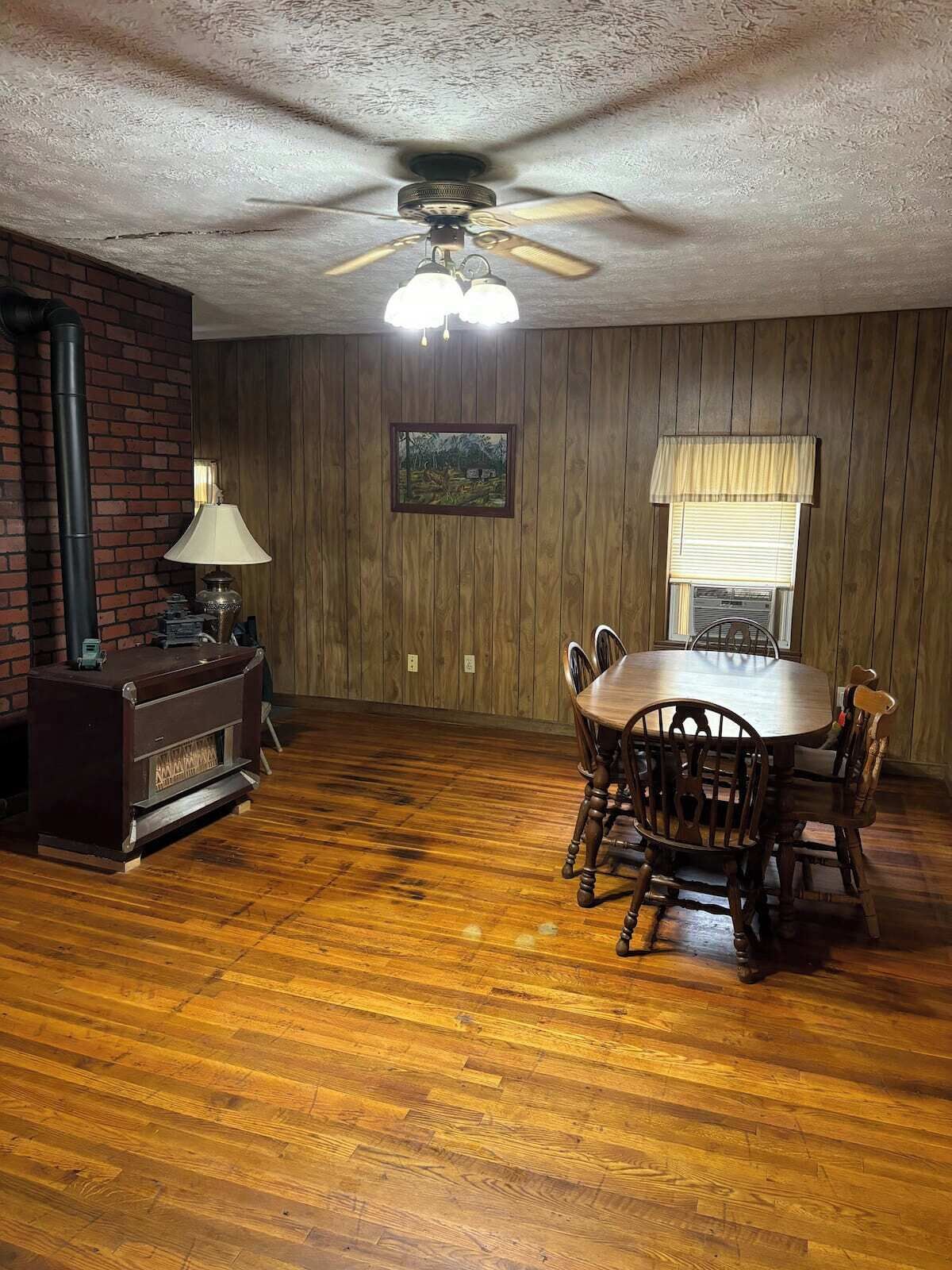 A dining room with a wooden table and chairs, a ceiling fan, a fireplace, and a window with blinds.