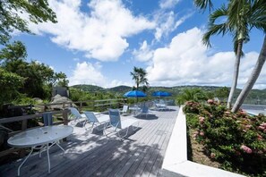 A wooden deck with lounge chairs and tables, surrounded by greenery and palm trees.
