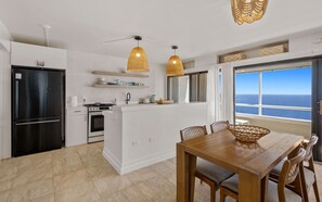 A modern kitchen with a central island, a dining area with a wooden table and chairs, and a view of the ocean through a large window.