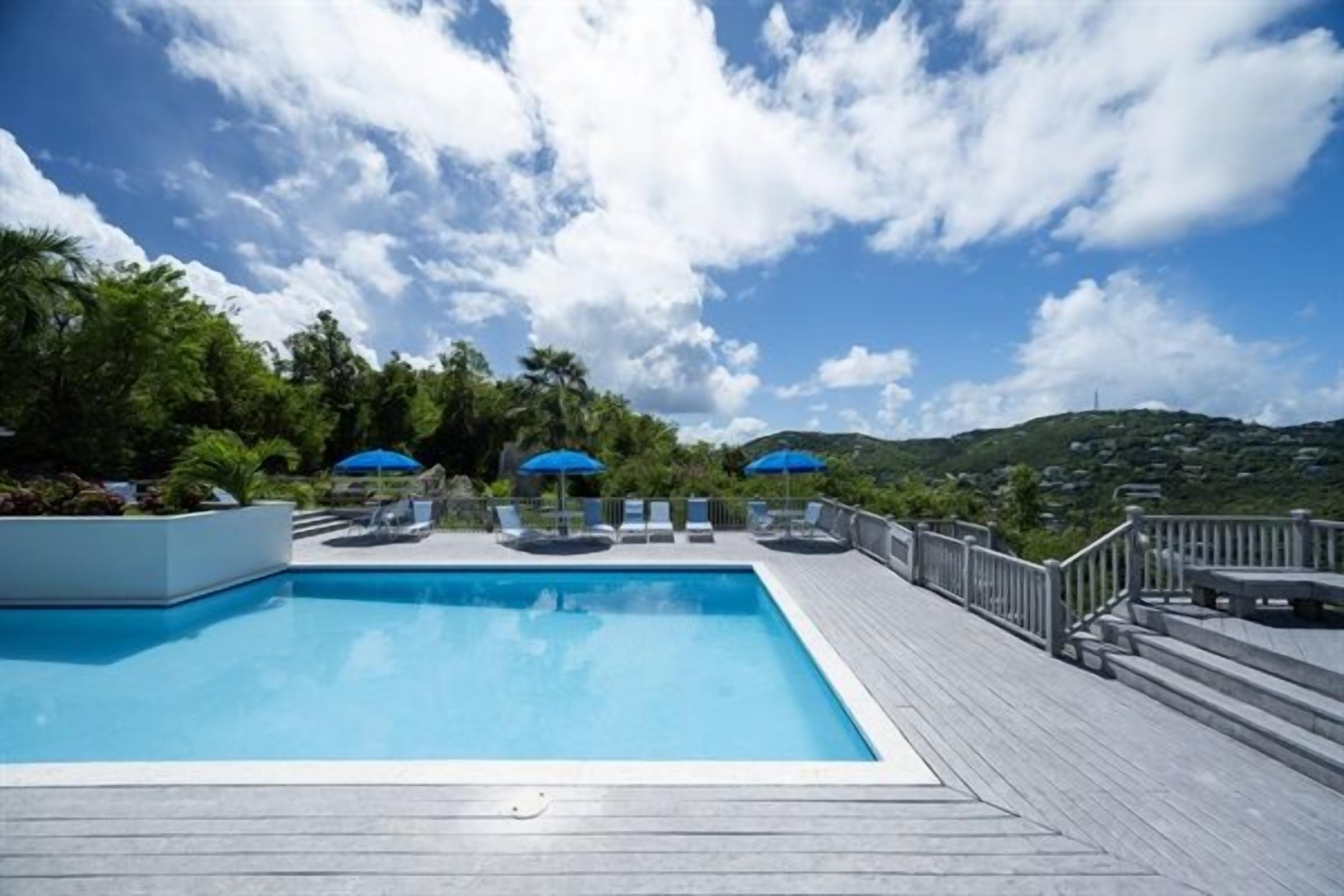 Ein Schwimmbecken mit Liegestühlen und Sonnenschirmen, umgeben von einer Holzterrasse und Grünpflanzen.