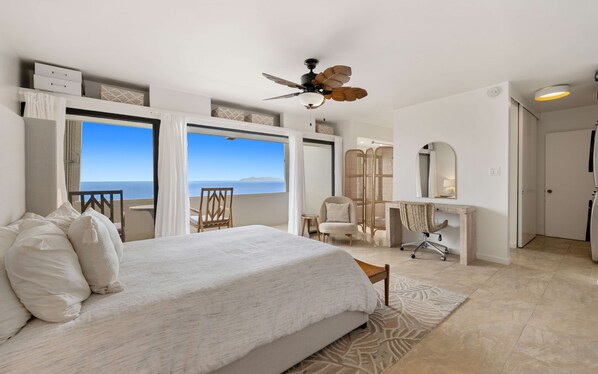 A hotel room with a bed, a desk, a chair, a ceiling fan, and a view of the sea.