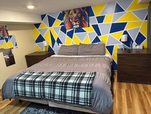 Une chambre moderne avec un mur orné d’un motif géométrique, un grand lit recouvert d’une couverture à carreaux et une table de chevet avec une lampe.