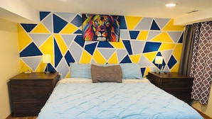 A bedroom with a geometric-patterned wall, a large bed with a blue quilt, two bedside tables with lamps, and a painting above the bed.