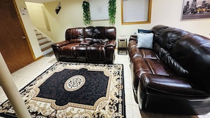 A living room with a brown leather sofa, a matching recliner, a patterned rug, a staircase, and a cityscape painting on the wall.