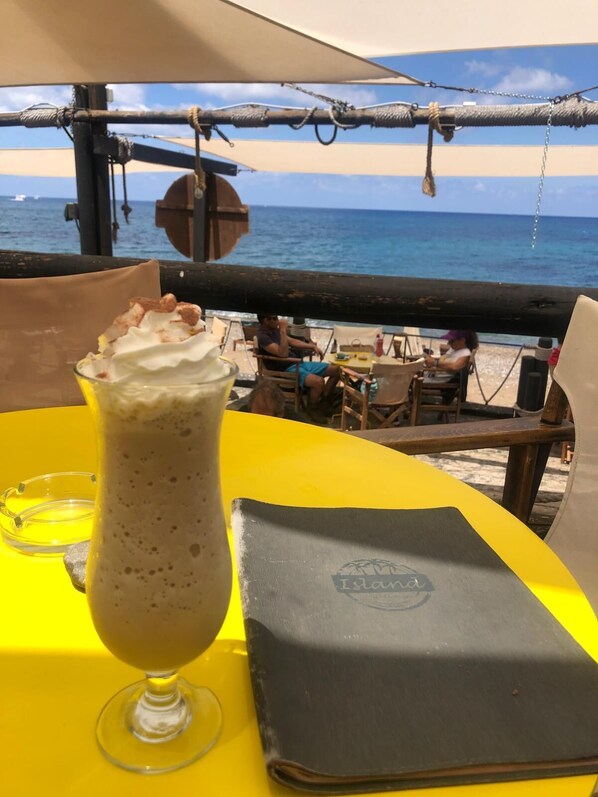 A table with a drink and a menu, overlooking a beach with people and a boat in the background. - Paphos Holiday Apartments Pool View Apartment (Paphos)