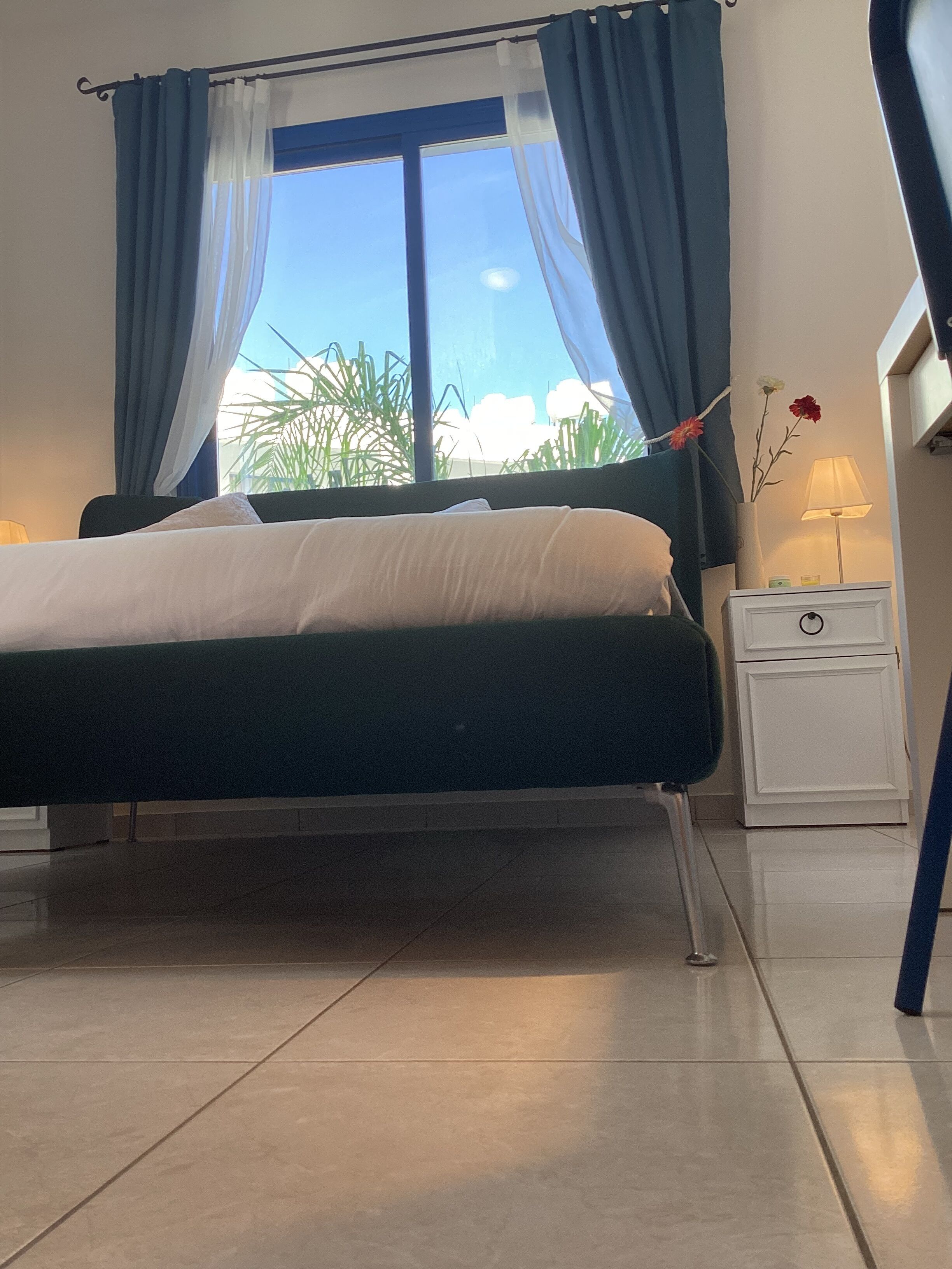 A bed with a white mattress and dark headboard, a nightstand with a lamp, and a window with blue curtains.