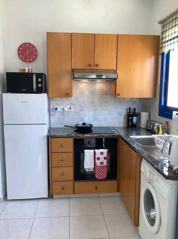 A compact kitchen with wooden cabinets, a built-in oven, a microwave, a sink, and a washing machine. - Paphos Holiday Apartments Pool View Apartment (Paphos)