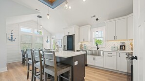 A modern kitchen with white cabinetry, a central island, and a dining area with wooden chairs. - Sandy Pelican: Pool, Hot Tub, Elevator, Close to Beach (Corolla)