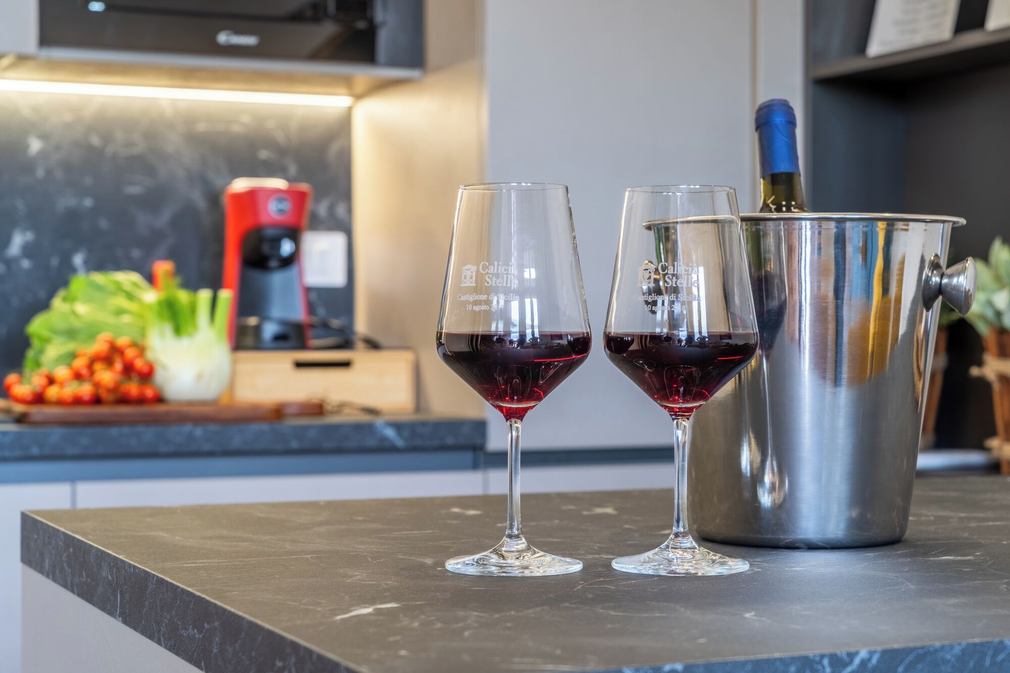 Two wine glasses filled with red wine on a kitchen counter next to a stainless steel wine bucket.