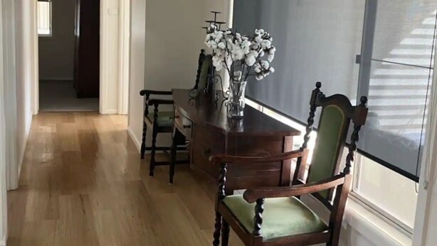 Un comedor con mesa y sillas de madera, un florero con flores y vistas a través de una ventana con persianas.