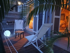 Eine Holzterrasse mit zwei weißen Stühlen und einem Tisch, umgeben von Palmenwedeln.
