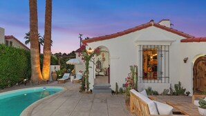 A white building with a red-tiled roof, a swimming pool, and a patio with wooden furniture and cacti. - Casa Gitana: Moroccan-Infused Spanish Retreat with Pool and Casita (Palm Springs)