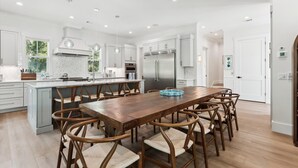 A modern kitchen with white cabinetry, a central island, and a dining area with wooden chairs. - 221 Cordillo at S Forest Beach, New Build, Easy to Beach, Private Pool (Hilton Head Island)