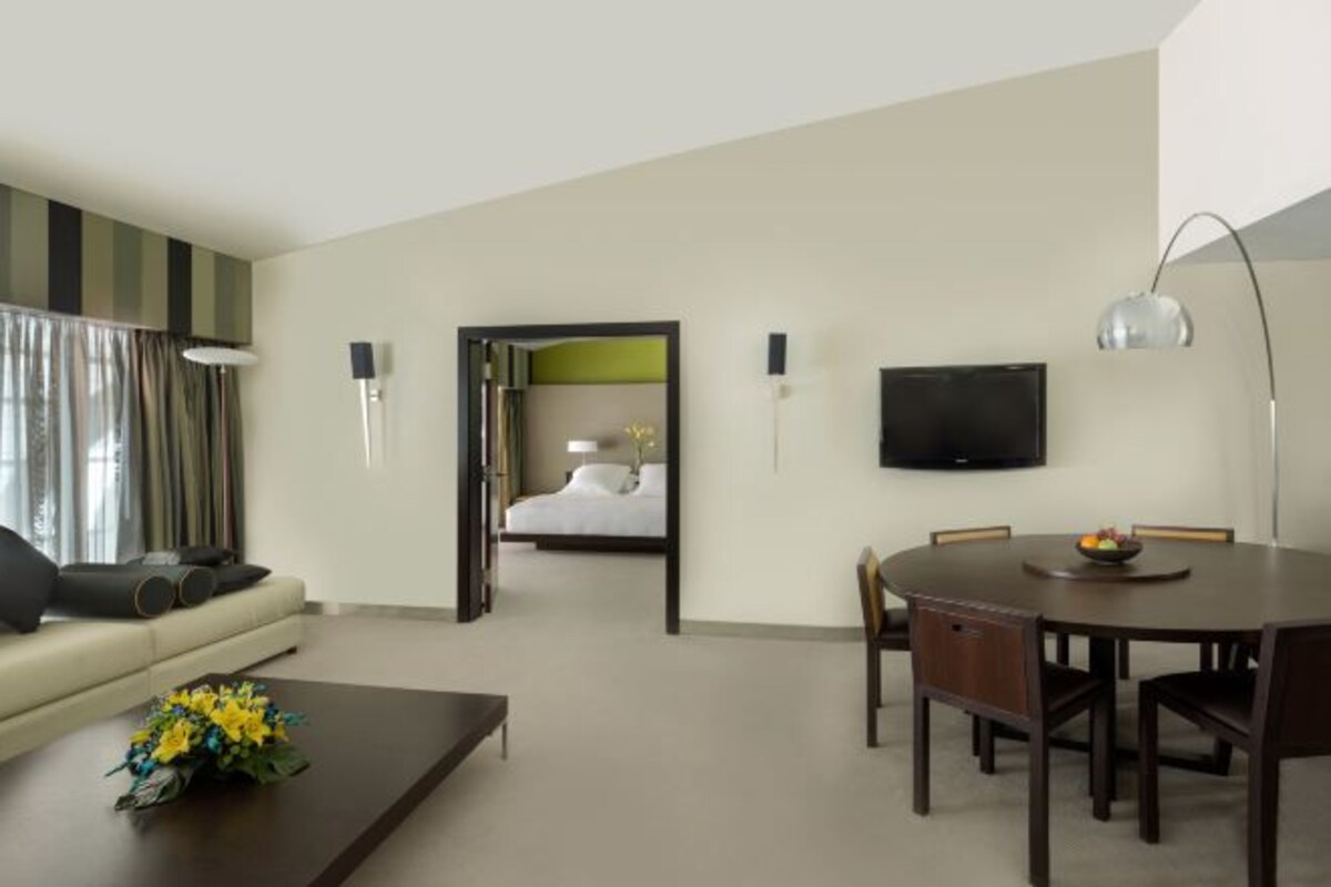Habitació d'hotel amb sofà, taula de cafè, taula de menjador, cadires, televisió de pantalla plana i llit amb capçalera verda.