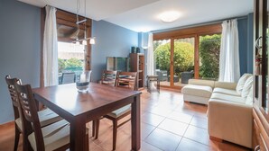 A dining area with a wooden table and chairs, a TV, and a sofa in a bright room with a view of greenery outside. - House Llanes (Poo) near the beach - Traudel (Llanes)