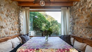 Una habitación con pared de piedra, techo de madera, mantel con motivos florales, un jarrón con flores y vistas al verdor exterior.