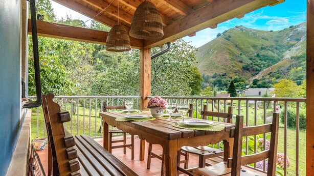 Una mesa de madera con platos, vasos y un jarrón en un balcón con vistas a colinas verdes.