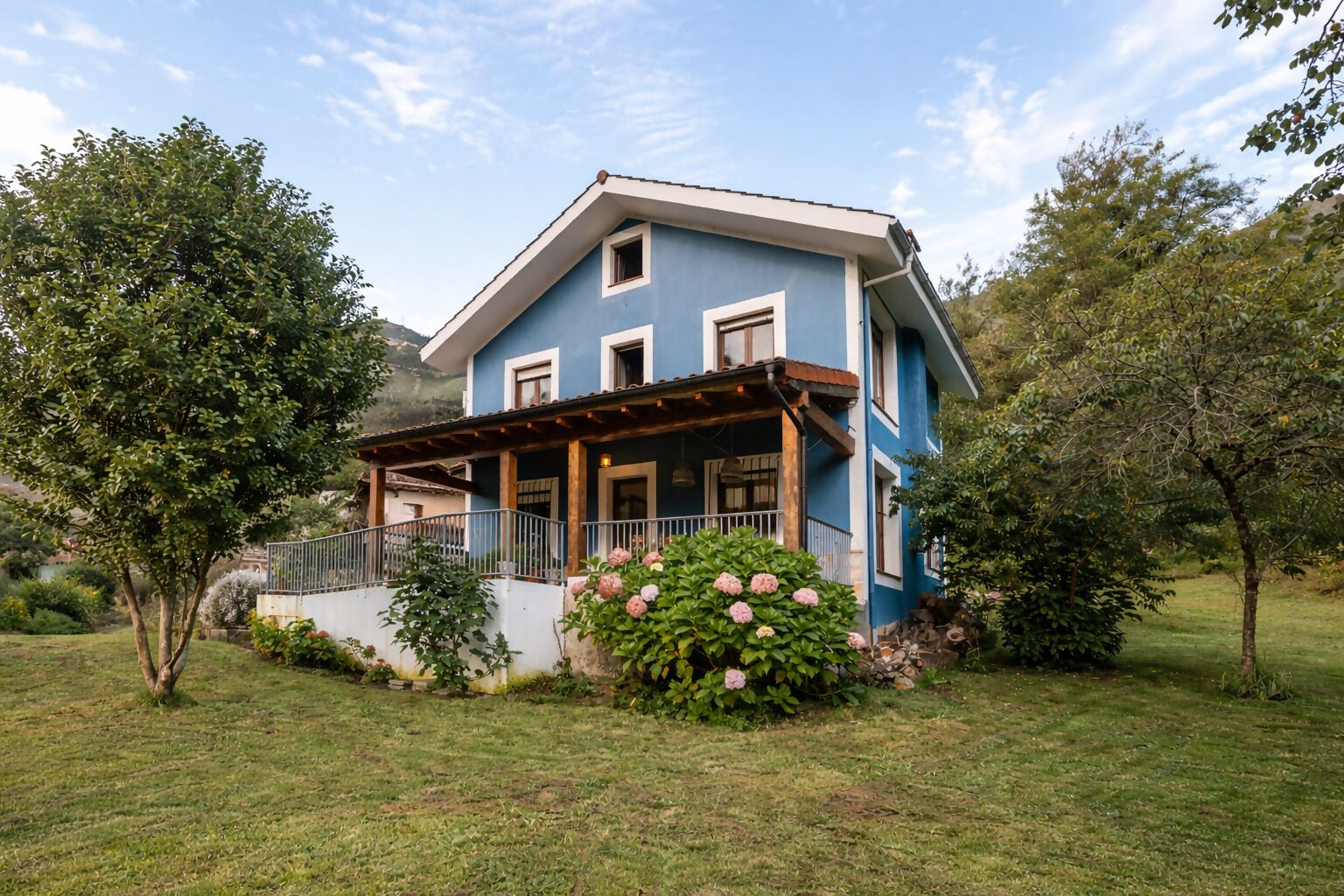 A two-story house with a blue exterior, a covered porch, and a garden with pink flowers.