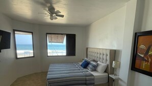 A bedroom with a bed, a ceiling fan, two windows with beach views, and a framed picture on the wall.
