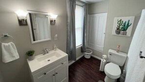 A bathroom with a white vanity, sink, and mirror. There is a toilet, a basket, and a cactus decoration on the wall.