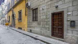 A narrow cobblestone street lined with historic buildings, featuring a yellow building with a statue and a brown door on a stone building. - Lietta Apartment in Florence (Firenze)