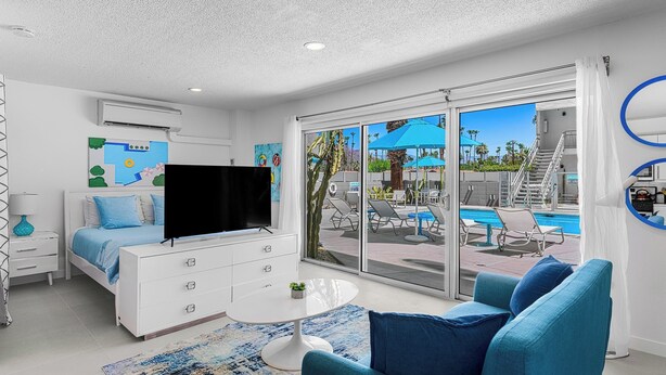A modern hotel room with a bed, a TV, a white dresser, a round table, and a view of a pool area. - South Palm Springs Studio - Unit 101 Float Apartments (Palm Springs)