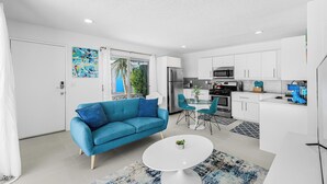 A modern living room with a blue sofa, a dining table, and a kitchen area with white cabinets and appliances. - South Palm Springs Studio - Unit 101 Float Apartments (Palm Springs)