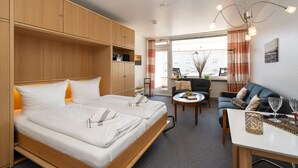 A modern hotel room with a large bed, a sofa, a dining table, and a view of the city. - Strandstraße - Cozy, central 1-room apartment, close to the beach, balcony, (Westerland)