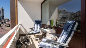 A balcony with two striped lounge chairs, a small table with a cup and saucer, and a view of a building with arched windows. - Strandstraße - Cozy, central 1-room apartment, close to the beach, balcony, (Westerland)