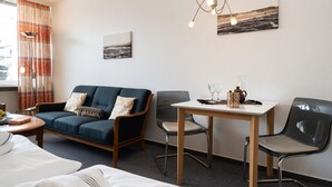 A hotel room with a sofa, two chairs, a small table with a teapot and glasses, a painting on the wall, and a window with curtains. - Strandstraße - Cozy, central 1-room apartment, close to the beach, balcony, (Westerland)