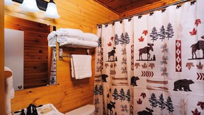 Ein Badezimmer einer Holzhütte mit Waschbecken, Toilette und Spiegel. Handtücher liegen auf einem Regal gestapelt und ein Duschvorhang mit Tiermustern ist angebracht.