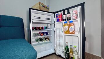Un refrigerador abierto repleto de diversas bebidas, incluyendo botellas y latas, y una caja de madera encima.