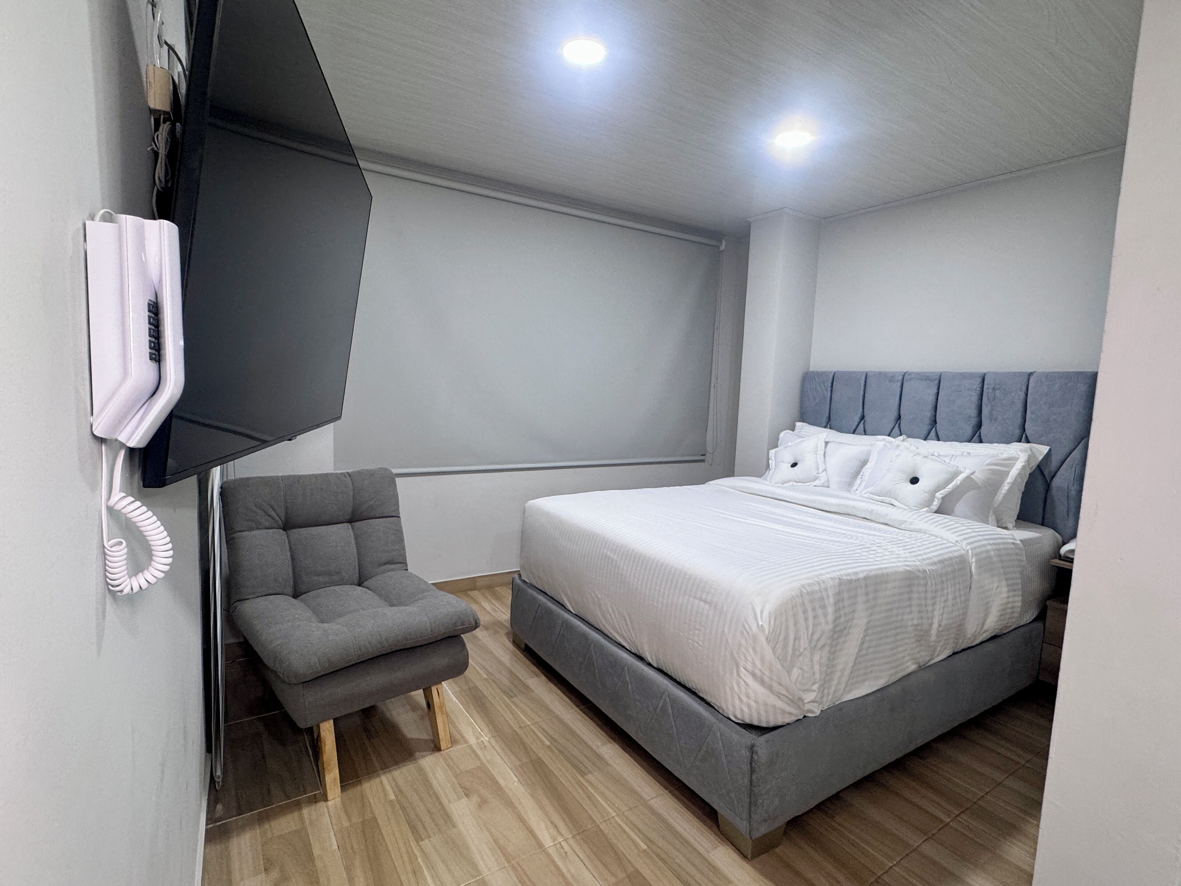Un dormitorio moderno con una butaca tapizada de gris, una cama con ropa de cama blanca y un televisor montado.