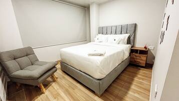 Un dormitorio moderno con una silla tapizada de gris, una cama con ropa blanca impecable y una mesita de noche de madera.