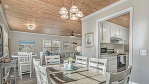 Un coin repas avec une table et des chaises en bois, un ventilateur de plafond et une cuisine visible à travers une porte ouverte.