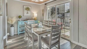 Un espace repas doté d’un plafond en bois, d’un lustre, d’une table avec des chaises, d’un buffet bleu et d’une grande porte-fenêtre coulissante donnant sur une terrasse.