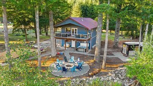 A two-story cabin with a balcony, surrounded by trees, featuring a fire pit and outdoor seating. - River Retreat w/ hot tub, beach & arcade/ game room (Packwood)
