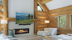 Interior de uma cabana de toras com lareira, uma TV montada e uma área de estar aconchegante.