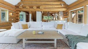 Interior de cabana de tronco com um sofá branco modular, uma mesa de centro de madeira e área de cozinha visível por uma porta aberta.