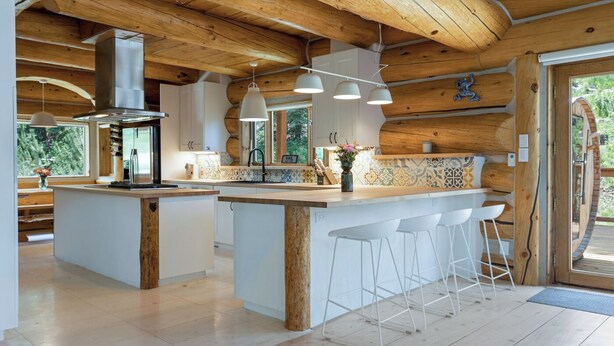 Uma cozinha moderna com vigas de madeira, uma ilha central com banquetas, luminárias pendentes e vista para o exterior.